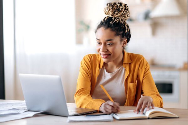 Focused cute stylish african american female student with afro dreadlocks, studying remotely from home, using a laptop, taking notes on notepad during online lesson, e-learning concept, smiling