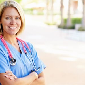 Smiling female nurse in blue scrubs standing outdoors with a stethoscope around her neck.