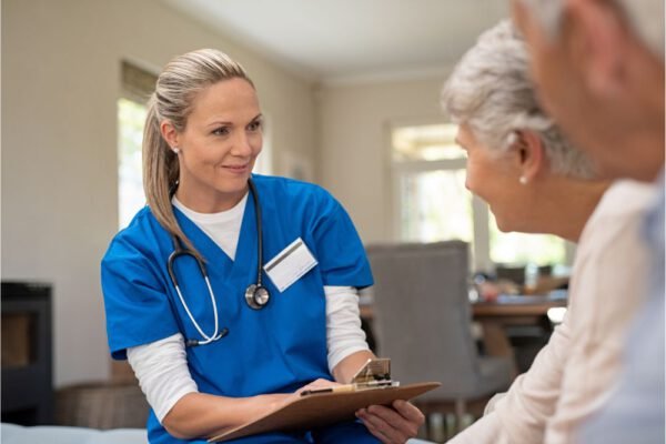 A cheerful elderly woman with short curly gray hair, dressed in a light blue shirt and gray cardigan, stands in a bright room, leaning on a cane. She looks fondly at a young female healthcare worker in a blue scrubs uniform, who has a stethoscope around her neck and an ID badge clipped to her pocket. The healthcare worker, with curly hair tied back and a warm smile, is gently placing her hand on the woman's arm in a comforting gesture. Behind them, the room is neatly decorated with framed artwork and has a welcoming atmosphere.