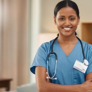 A cheerful elderly woman with short curly gray hair, dressed in a light blue shirt and gray cardigan, stands in a bright room, leaning on a cane. She looks fondly at a young female healthcare worker in a blue scrubs uniform, who has a stethoscope around her neck and an ID badge clipped to her pocket. The healthcare worker, with curly hair tied back and a warm smile, is gently placing her hand on the woman's arm in a comforting gesture. Behind them, the room is neatly decorated with framed artwork and has a welcoming atmosphere.
