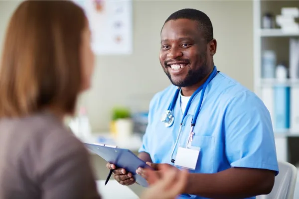 Black Male nurse in blue scrubs holding a clip board. Talking to a patient.