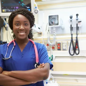 Black nurse standing with her arms crossed. Wearing a blue scrub top in a hospital room.