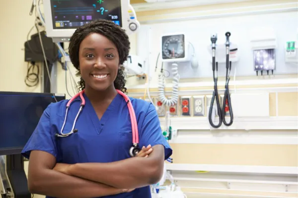 Black nurse standing with her arms crossed. Wearing a blue scrub top in a hospital room.