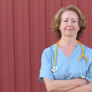 White female nurse wearing a blue scrub top. Standing next to a metal shed.