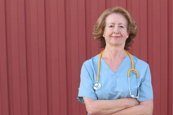 White female nurse wearing a blue scrub top. Standing next to a metal shed.