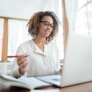 A smiling woman doing a PN Remediation with curly hair and glasses is focused on her work in a bright, naturally lit room. She wears a white blouse and is holding a pen, suggesting she's either taking notes or about to annotate a document.