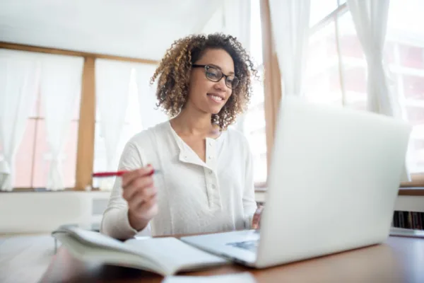 A smiling woman doing a PN Remediation with curly hair and glasses is focused on her work in a bright, naturally lit room. She wears a white blouse and is holding a pen, suggesting she's either taking notes or about to annotate a document.